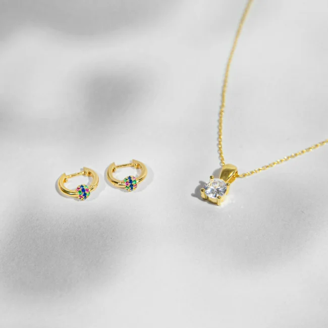 Sidabriniai papuošalai: auskarai, ir pakabukas. Šis rinkinys, sudarytas iš auksu dengtų žiedo formos auskarų ir žavingo kaklo pakabuko, yra tikras džiaugsmo ir spalvų šventimas. Auskaruose išsidėstę spalvingi kubiniai cirkoniai suteikia žaismingą ir energingą išvaizdą, o kaklo pakabuko centrinę vietą užima didelis, švytintis kubinis cirkonis, kuris yra tikras žvilgsnio traukos centras. Komplektas puikiai tinka tiems, kurie mėgsta pridėti šiek tiek spalvos prie savo kasdieninio ar vakarinio įvaizdžio, nes jis neabejotinai pritrauks dėmesį ir pagyvins bet kokią aprangą.